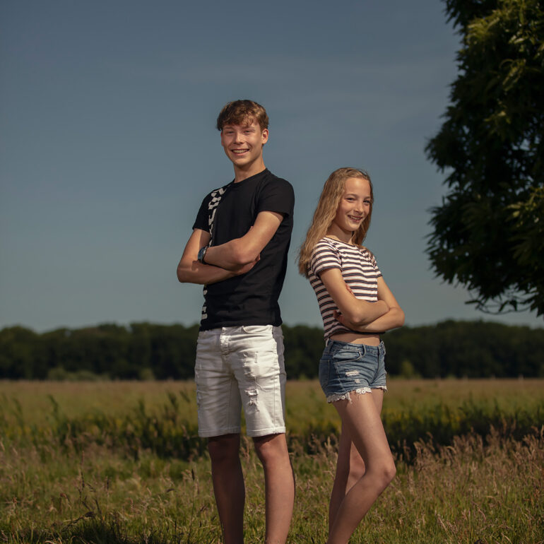Jongen en meisje staan met gekruiste armen bij een grasveld onder een blauwe lucht.