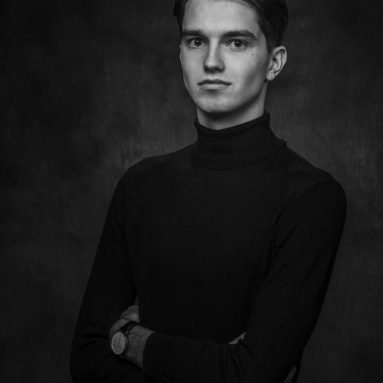 Zwart-witportret van een jonge man in een donkere coltrui, met gekruiste armen tegen een donkere achtergrond.
