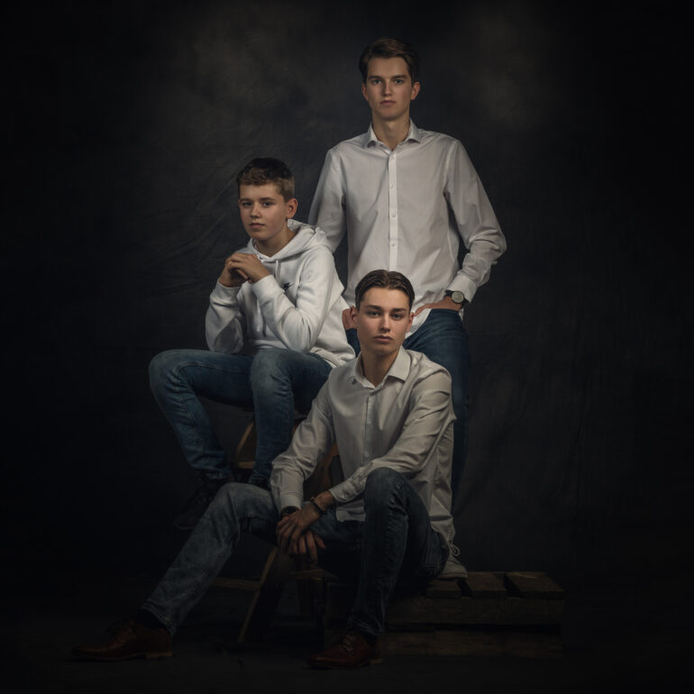Drie jongens in lichte overhemden en een hoodie zitten en staan bij houten kratten tegen een donkere achtergrond.