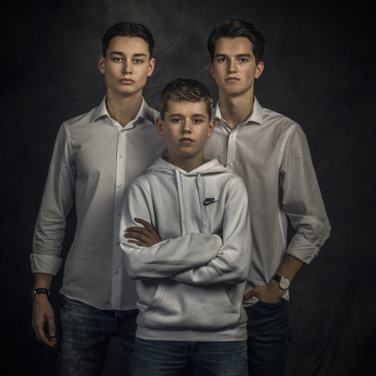 Drie jongens in lichte kleding staan dicht bij elkaar tegen een donkere achtergrond.
