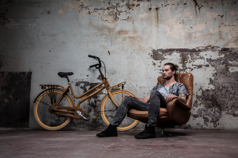Man met tatoeages zit in een bruine leren fauteuil naast een goudbruine Batavus-fiets voor een verweerde muur.