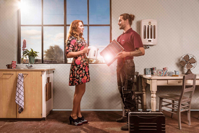 Vrouw met een witte kan praat met een monteur die een metalen onderdeel vasthoudt in een keuken met retro inrichting.