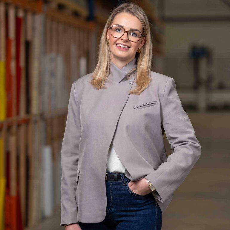Vrouw met bril en een lichtgrijs colbert staat voor een onscherpe bedrijfsachtergrond.