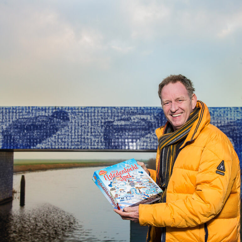 Piet Paulusma in een gele jas houdt een boek vast bij een brug over het water.
