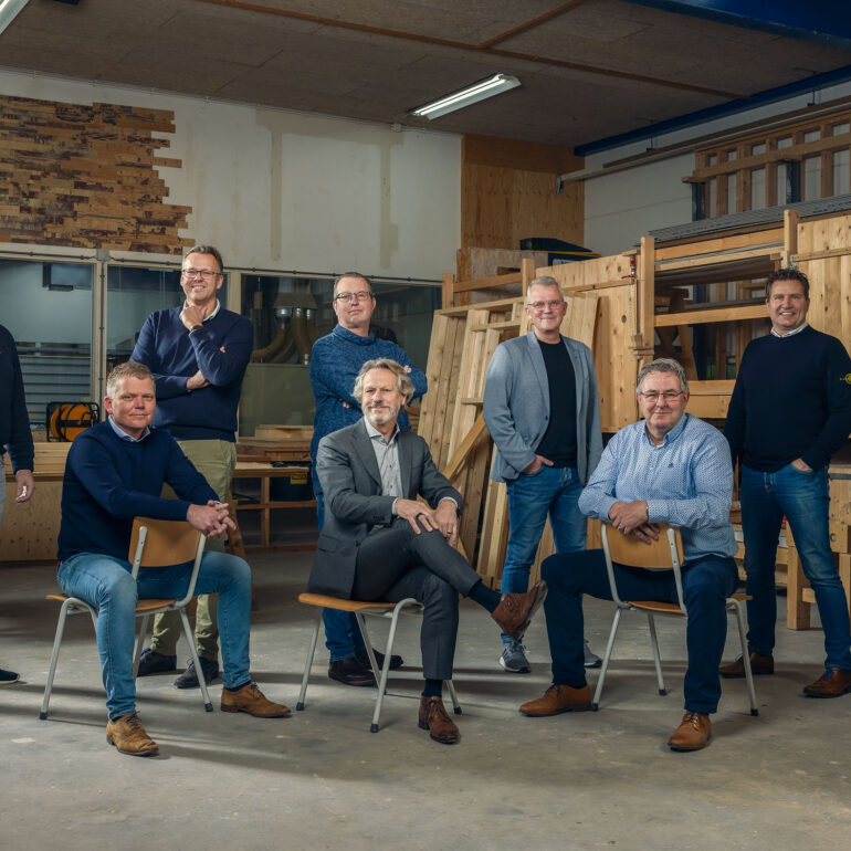 Zeven mannen poseren op en rond stoelen in een werkplaats met houten materialen.