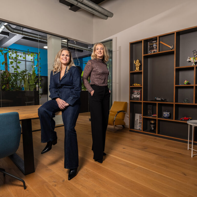 Twee vrouwen in zakelijke kleding poseren in een kantoor met een houten vloer en open kasten.