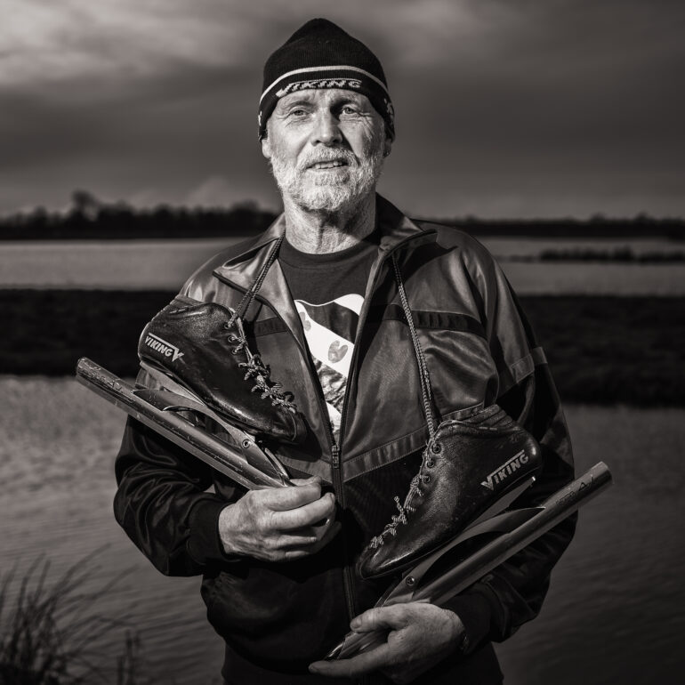 Zwart-witportret van een oudere man met baard en muts, die lange schaatsen vasthoudt bij het water.