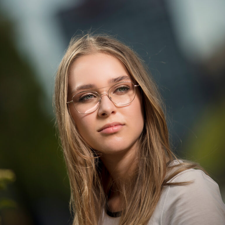 Jonge vrouw met lang haar en een bril kijkt naar de camera voor een onscherpe groene achtergrond.