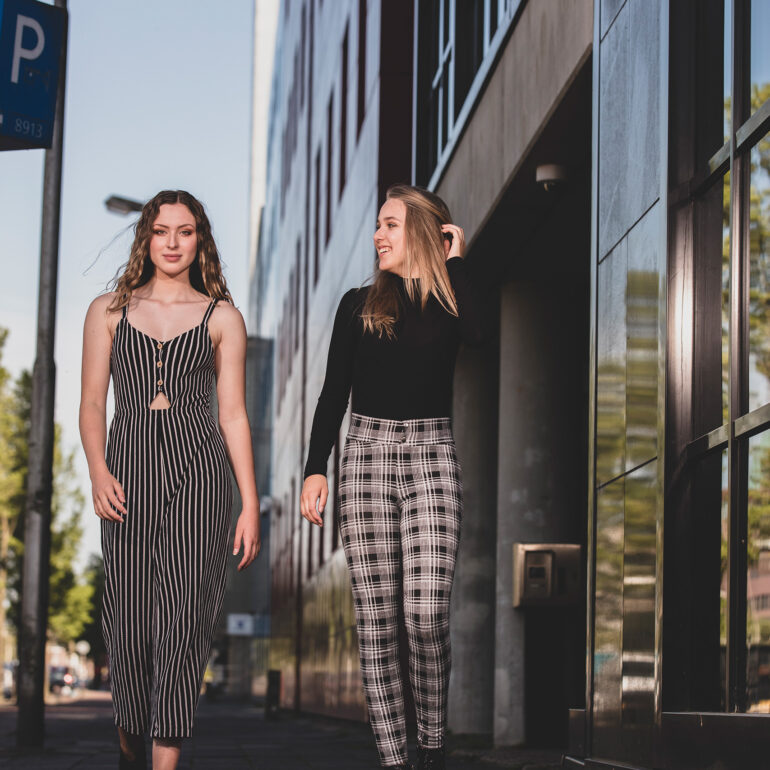 Twee jonge vrouwen staan naast elkaar bij de gevel van een modern gebouw, in een gestreepte jumpsuit en een geruite broek.