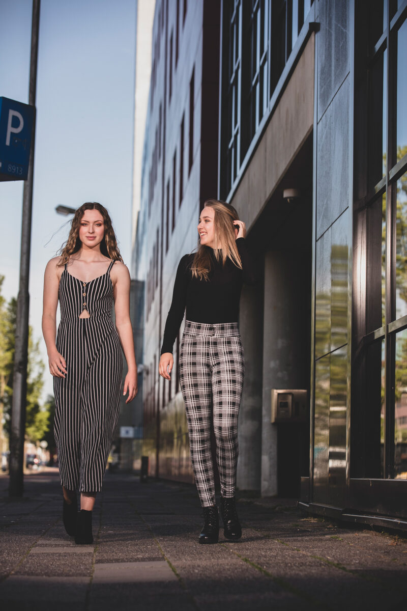Twee jonge vrouwen staan naast elkaar bij de gevel van een modern gebouw, in een gestreepte jumpsuit en een geruite broek.