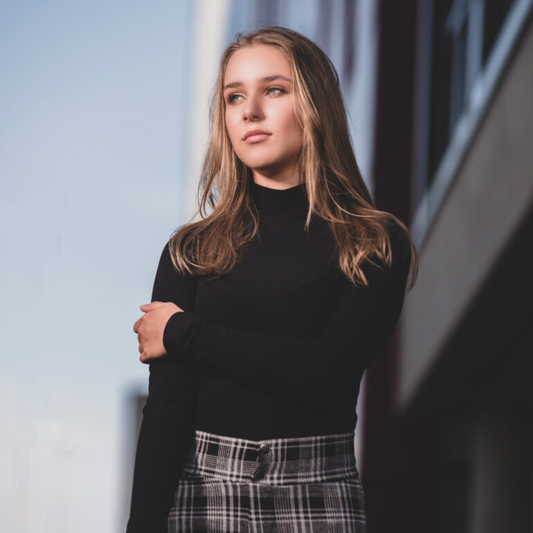 Jonge vrouw met lang haar, een zwarte coltrui en een geruite broek leunt bij een moderne gevel.