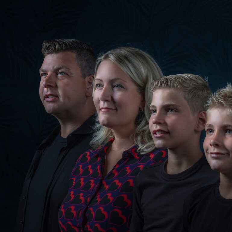 Twee volwassenen en twee jongens staan dicht bij elkaar en kijken dezelfde kant op tegen een donkere achtergrond.