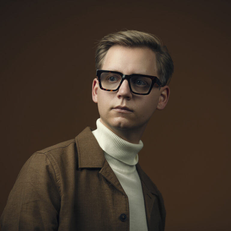 Jongen met een grote donkere bril, een lichte coltrui en een bruin jasje tegen een bruine achtergrond.