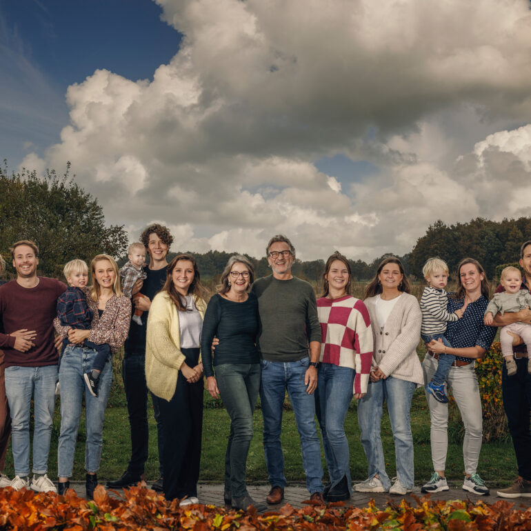 Grote familie staat buiten bij elkaar, met herfstbladeren op de voorgrond en een wolkenlucht boven het landschap.