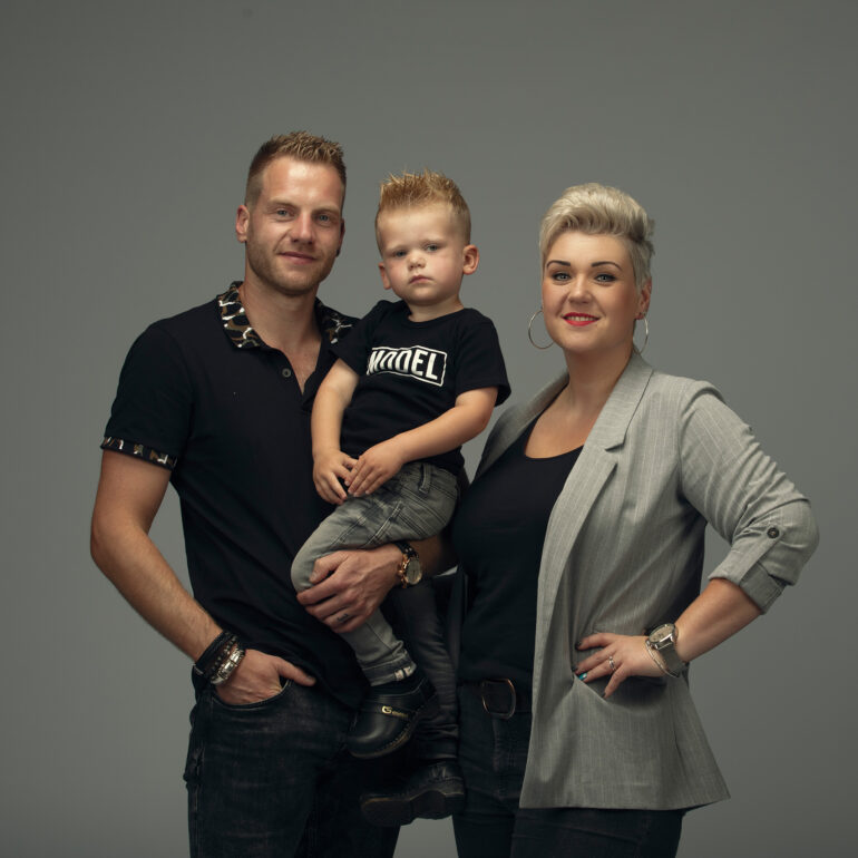 Man en vrouw poseren met een jong kind dat door de man wordt vastgehouden, tegen een grijze achtergrond.