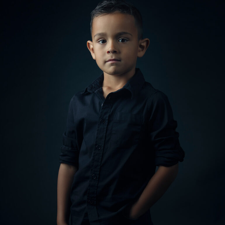 Jongen met kort donker haar en een donker overhemd staat met zijn handen in zijn zakken tegen een donkere achtergrond.