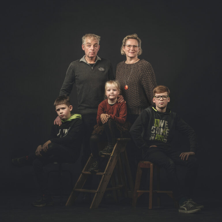 Twee volwassenen en drie kinderen poseren bij een houten ladder, een kruk en een stoel tegen een donkere achtergrond.