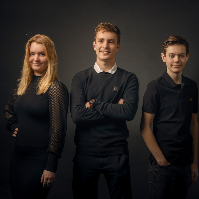 Jonge vrouw en twee jonge mannen in donkere kleding staan naast elkaar tegen een donkere achtergrond.