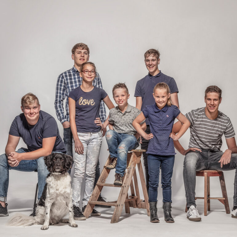 Groep kinderen en jongeren poseert met een hond op en rond krukken en een houten trapje tegen een lichte achtergrond.