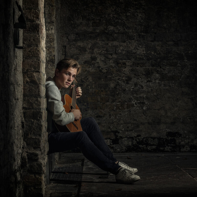 Jongen met een gitaar zit in een nis tussen oude stenen muren.