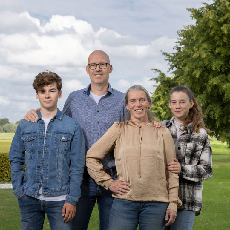 Twee volwassenen en twee kinderen staan dicht bij elkaar op het gras, met bomen en wolken op de achtergrond.