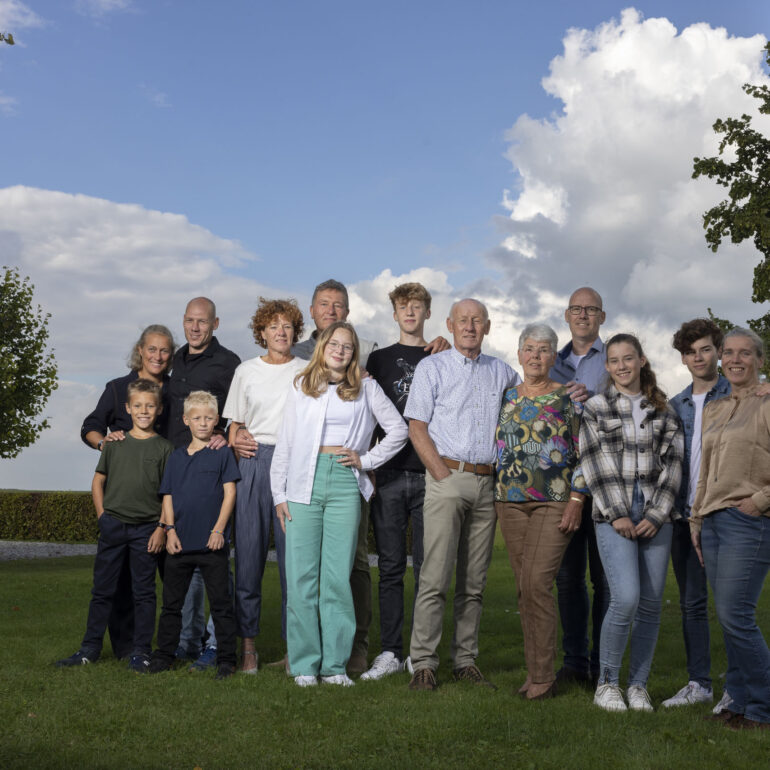 Grote familie met volwassenen en kinderen staat op een grasveld onder een blauwe lucht met wolken.