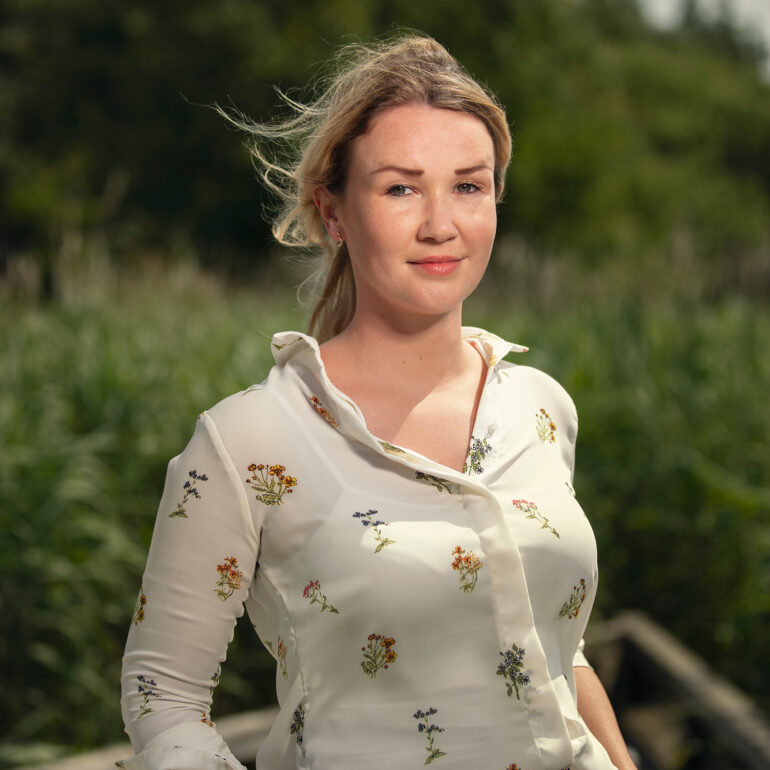 Jonge vrouw in een lichte blouse met bloemenprint staat buiten voor een groene achtergrond.