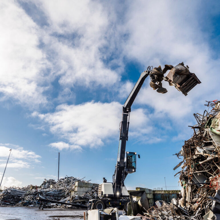 Grijperkraan tilt een metalen voorwerp boven een grote berg schroot op een recyclingterrein