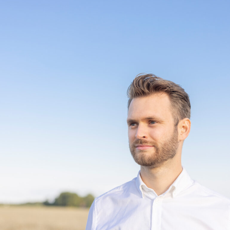Man met kort haar, een baard en een wit overhemd staat buiten bij een open veld onder een blauwe lucht.