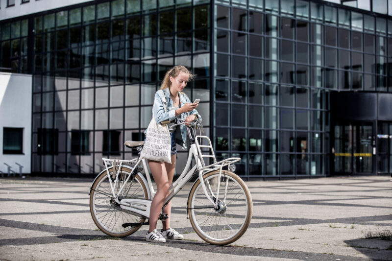 Jonge vrouw in een spijkerjas staat naast een witte stadsfiets en kijkt op haar telefoon voor een glazen gebouw.