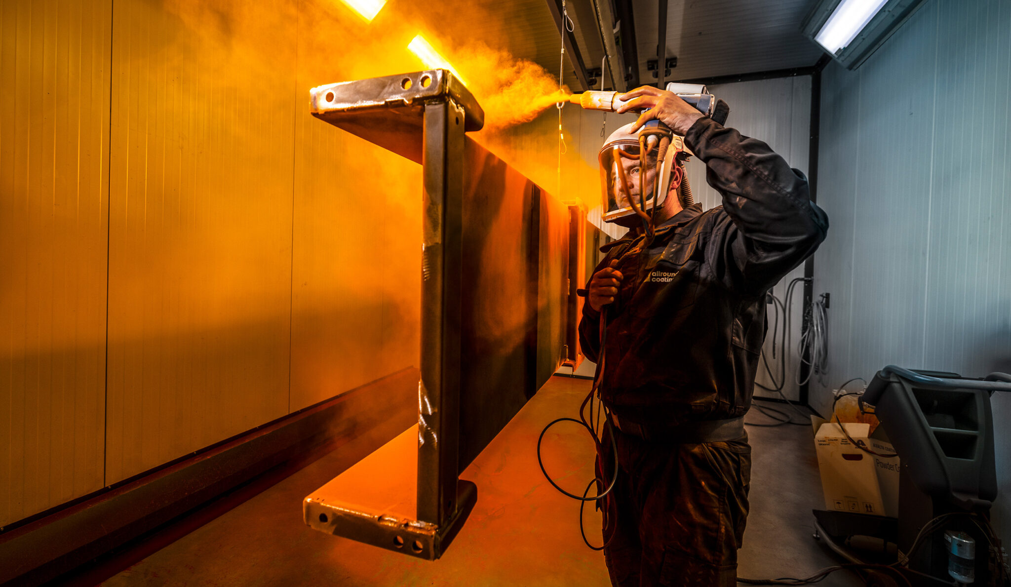 Medewerker spuit een hangend metalen onderdeel in een ruimte met warm oranje en koel blauw licht.