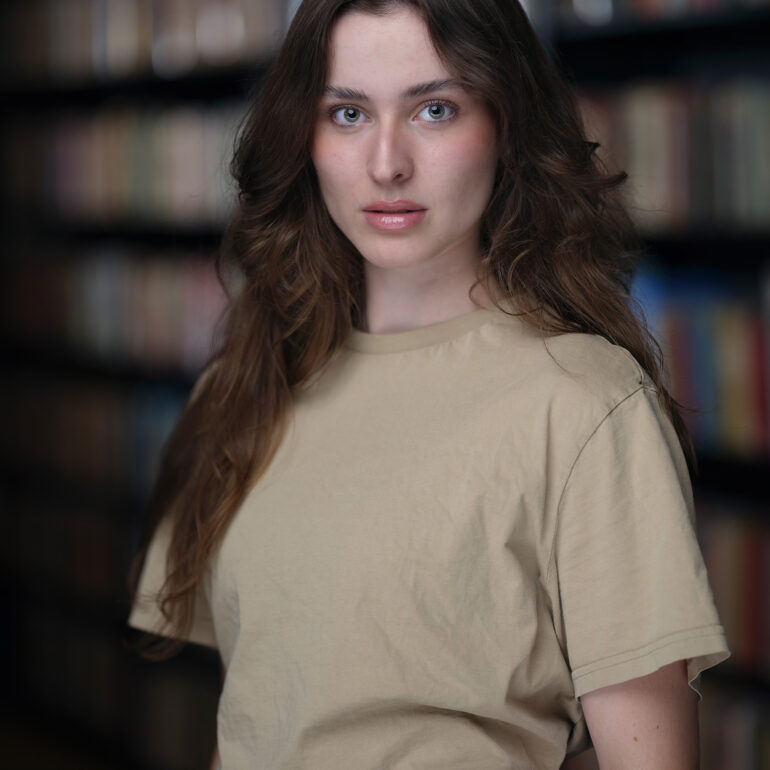 Jonge vrouw met lang bruin haar en een beige T-shirt voor een onscherpe boekenkast.