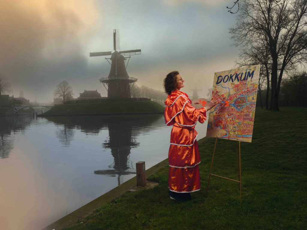 Personage in oranje kleding schildert op een plattegrond van Dokkum, met water en een molen op de achtergrond.
