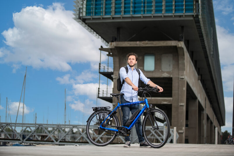 Man met een rugzak staat met een blauwe Batavus-fiets bij het water voor een modern gebouw.