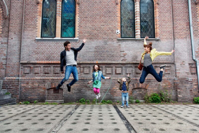 Twee volwassenen en een meisje springen naast een kleine jongen voor een bakstenen gevel met hoge ramen; boven hen hangt een voetbal.