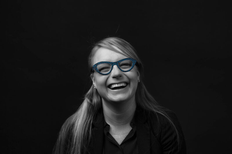 Lachende vrouw draagt een blauw cat-eye-brilmontuur in een verder zwart-witportret tegen een donkere achtergrond.