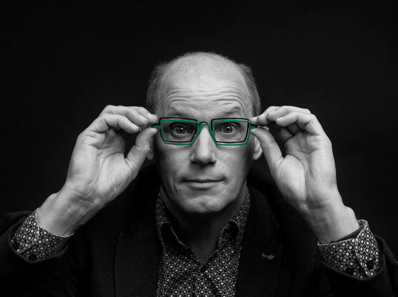 Man houdt met beide handen een groen rechthoekig brilmontuur vast in een verder zwart-witportret.