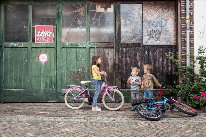 Drie kinderen staan met roze, blauwe en rode kinderfietsen voor verweerde groene deuren.