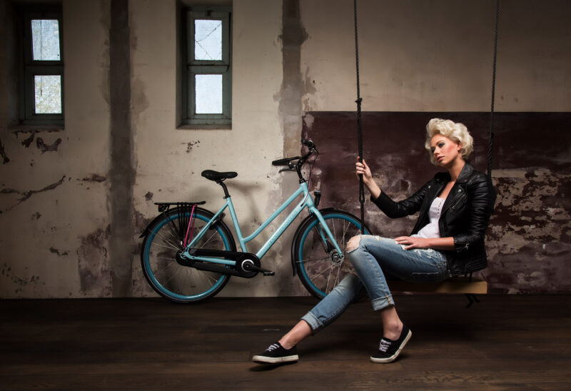 Blonde vrouw in een leren jas zit op een schommel naast een turquoise Batavus-fiets in een industriële ruimte.