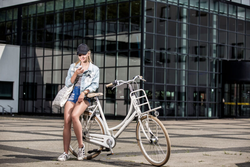Jonge vrouw met een pet zit op een witte stadsfiets en kijkt op haar telefoon voor een glazen gevel.