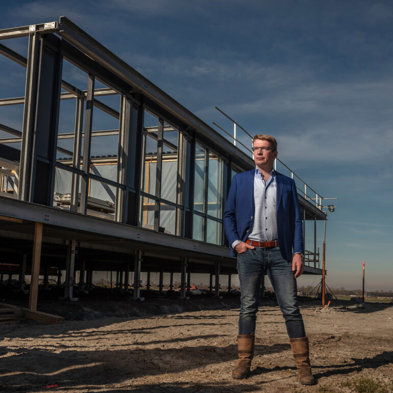 Man met een bril, blauw colbert en bruine laarzen staat op een bouwterrein voor een stalen gebouwconstructie.