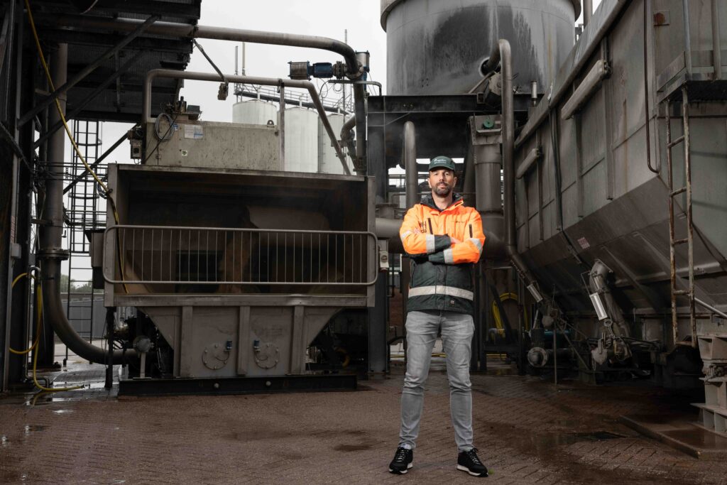 Agrifirm-medewerker in oranje veiligheidskleding tussen industriële installaties.