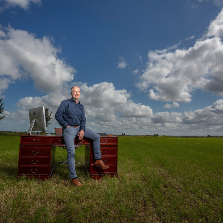 Man in een blauw overhemd zit op een houten bureau met een computer, midden in een groen weiland onder een wolkenlucht.