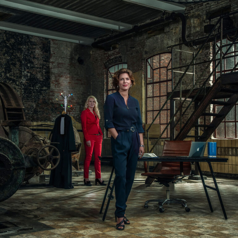Twee vrouwen in blauwe en rode zakelijke kleding poseren bij een bureau en een metalen trap in een oud fabriekspand.